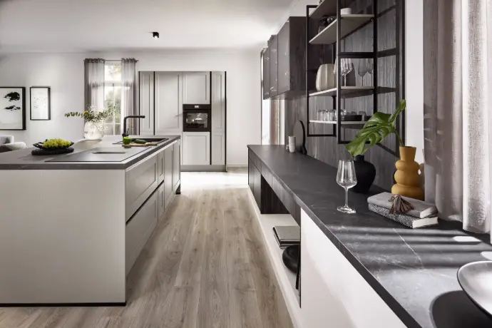 Moderne Landhausküche mit großer Kücheninsel, dunkler Arbeitsplatte, offenen Regalen und Glasvitrine. Lichtdurchfluteter Raum mit warmem Holzboden und stilvoller Dekoration.