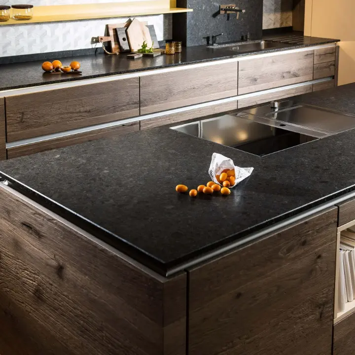 Dunkle Keramikarbeitsplatte auf warmen Holzfronten in einer modernen Küche, mit integrierter Kochfläche und dezentem Licht – Planung von LS Küchen Wohnen & Service.