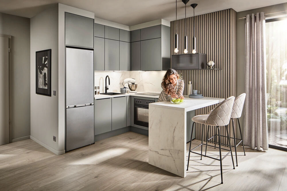 Moderne Wohnküche mit Kochinsel und Barhocker – offene Raumgestaltung für geselliges Kochen und Leben. Planung von LS Küchen. Moderne Wohnküche im Alltag: Frau nutzt Marmor-Kochinsel mit Barhocker – offene Raumgestaltung mit Holzelementen