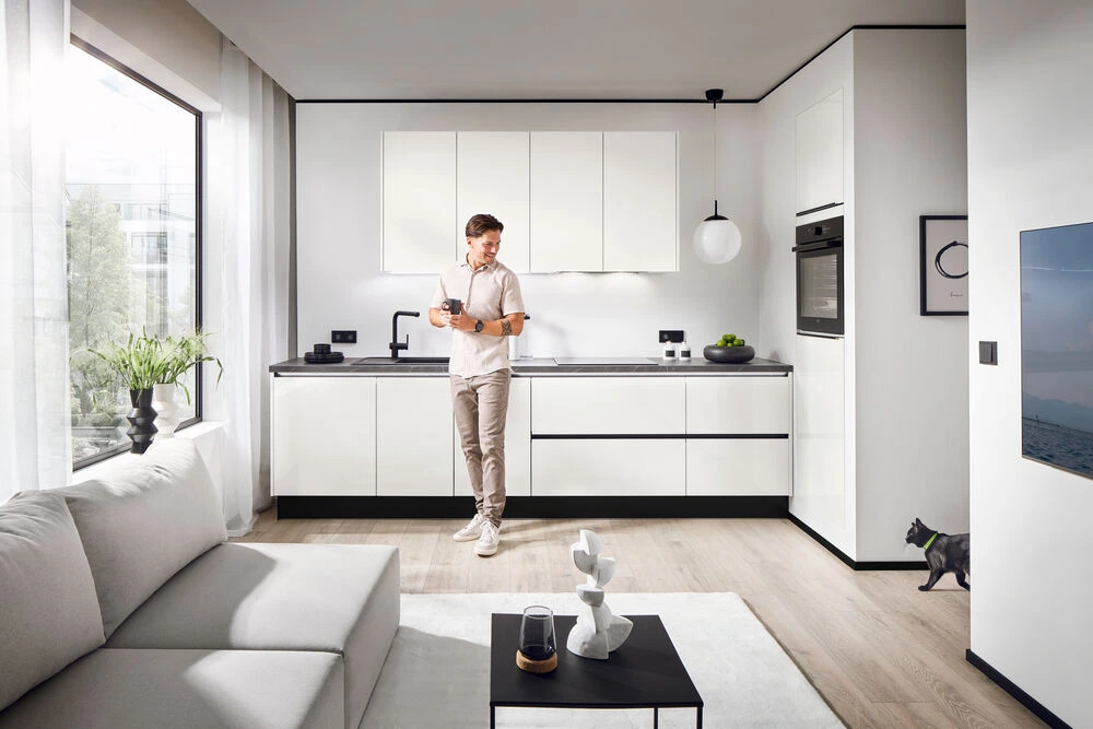 Das ist Wohnküche: Vom Sofa aus sehen, wer gerade kocht. Zusammen sein, auch wenn jemand am Herd steht. Person an Arbeitsplatte in offener Wohnküche: Alltagsnutzung mit Sichtverbindung zum Wohnzimmer – weisse Fronten
